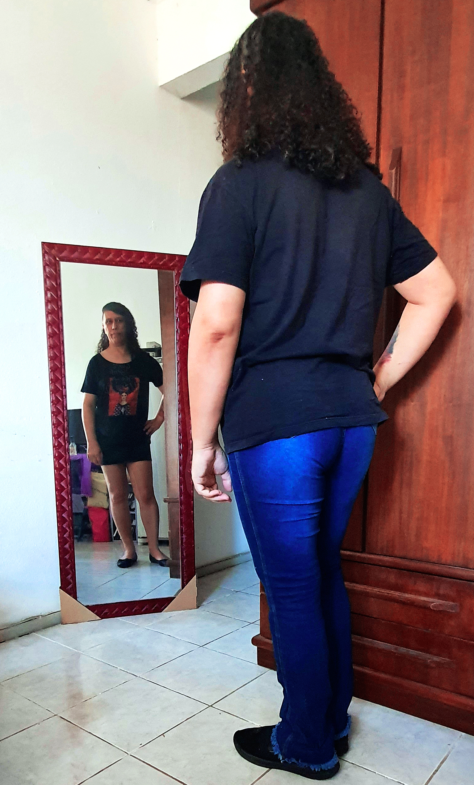 Pessoa se olhando no espelho, usando camisa preta, calça jeans azul e tênis. O reflexo no espelho está usando blusa preta, minissaia preta e sapatilhas.