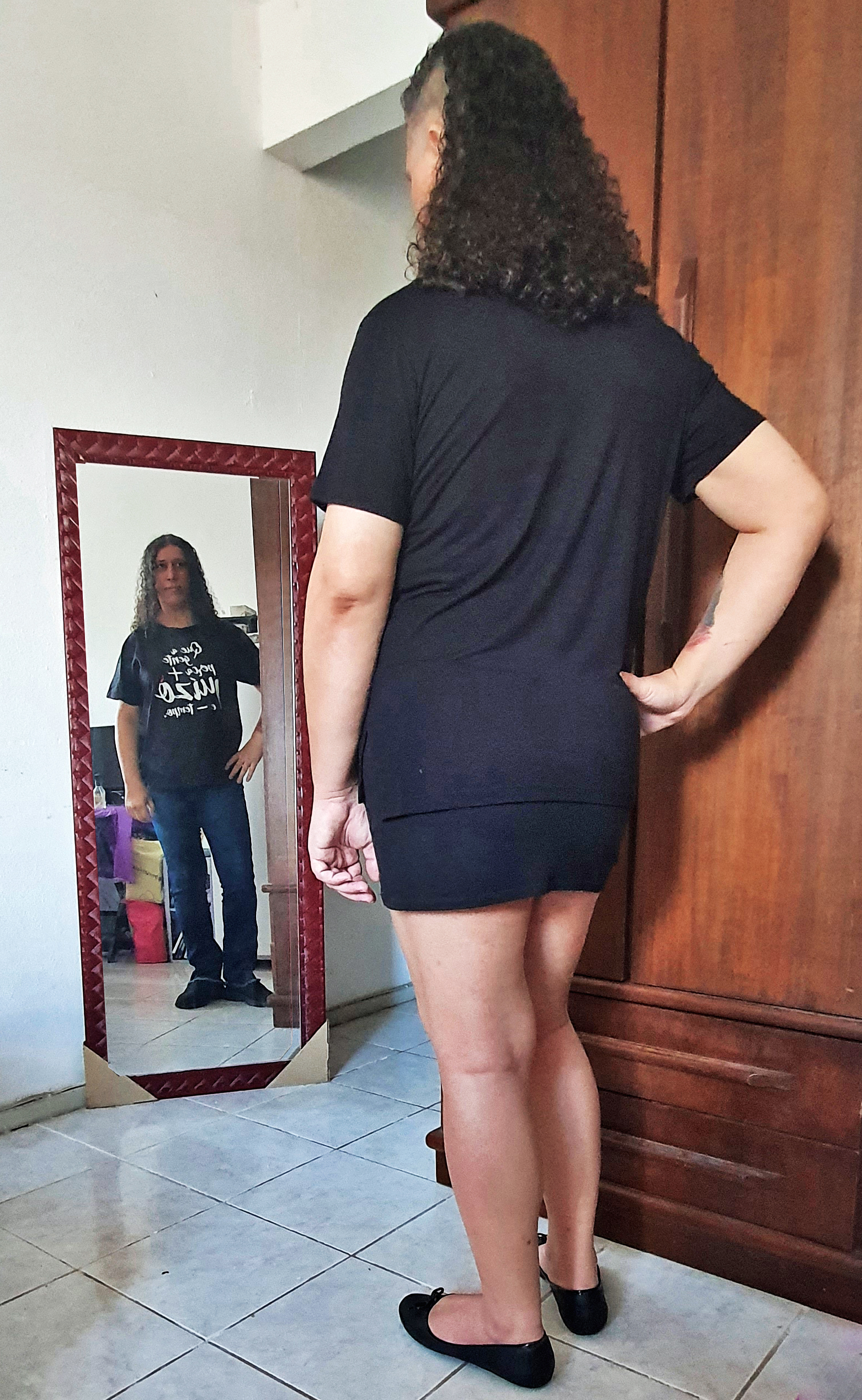 Pessoa se olhando no espelho, usando blusa preta, minissaia preta e sapatilhas. O reflexo está usando camisa preta, calça jeans azul e tênis.